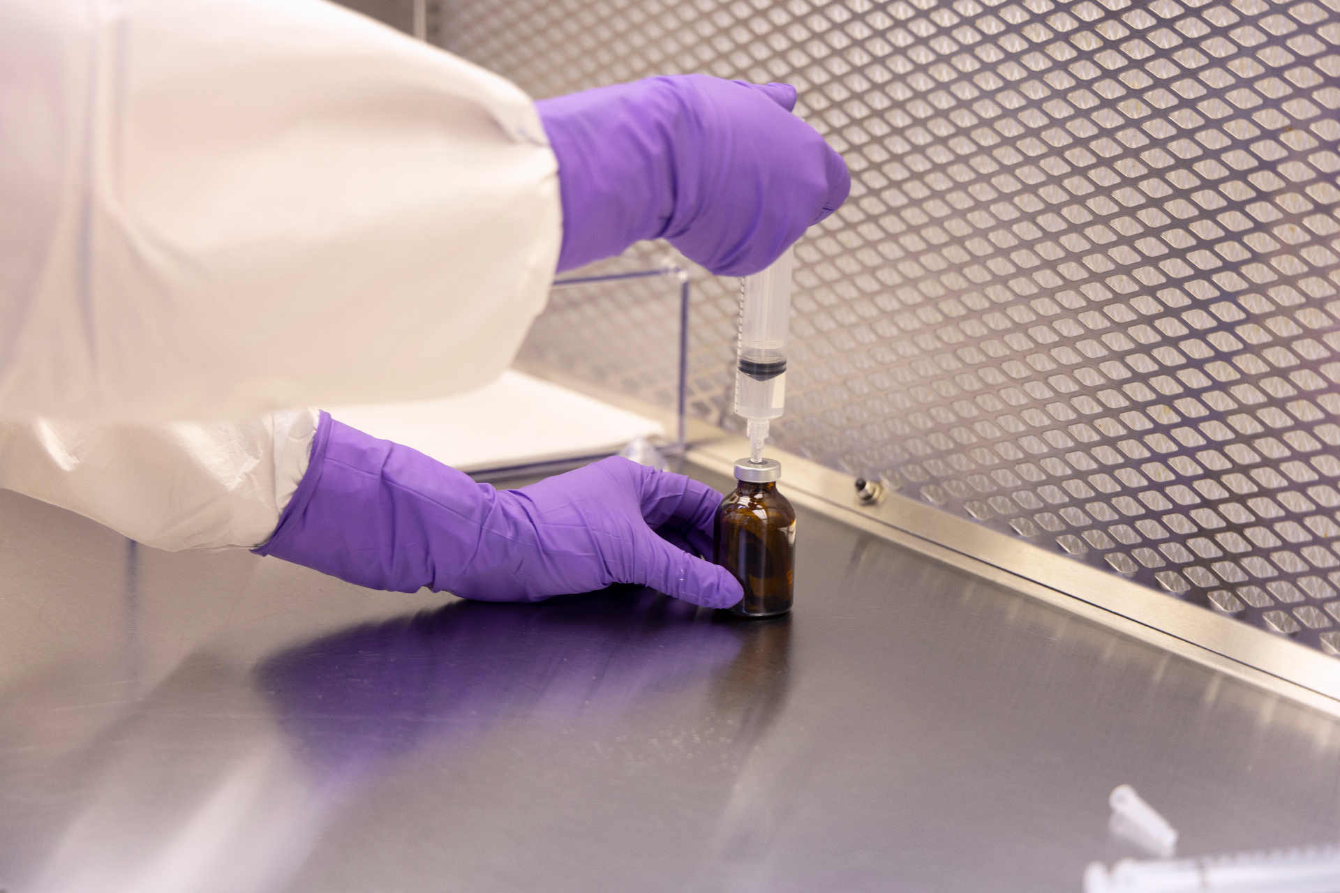 Pharmacist in purple gloves compounding a sterile ophthalmic vial in a cleanroom hood